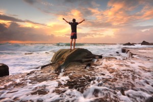 Teen boy stands on a rock among turbulent ocean seas and fast flowing water at sunrise. Worship praise zest adenture solitude finding peace among lifes turbulent times. Overcoming adversity.